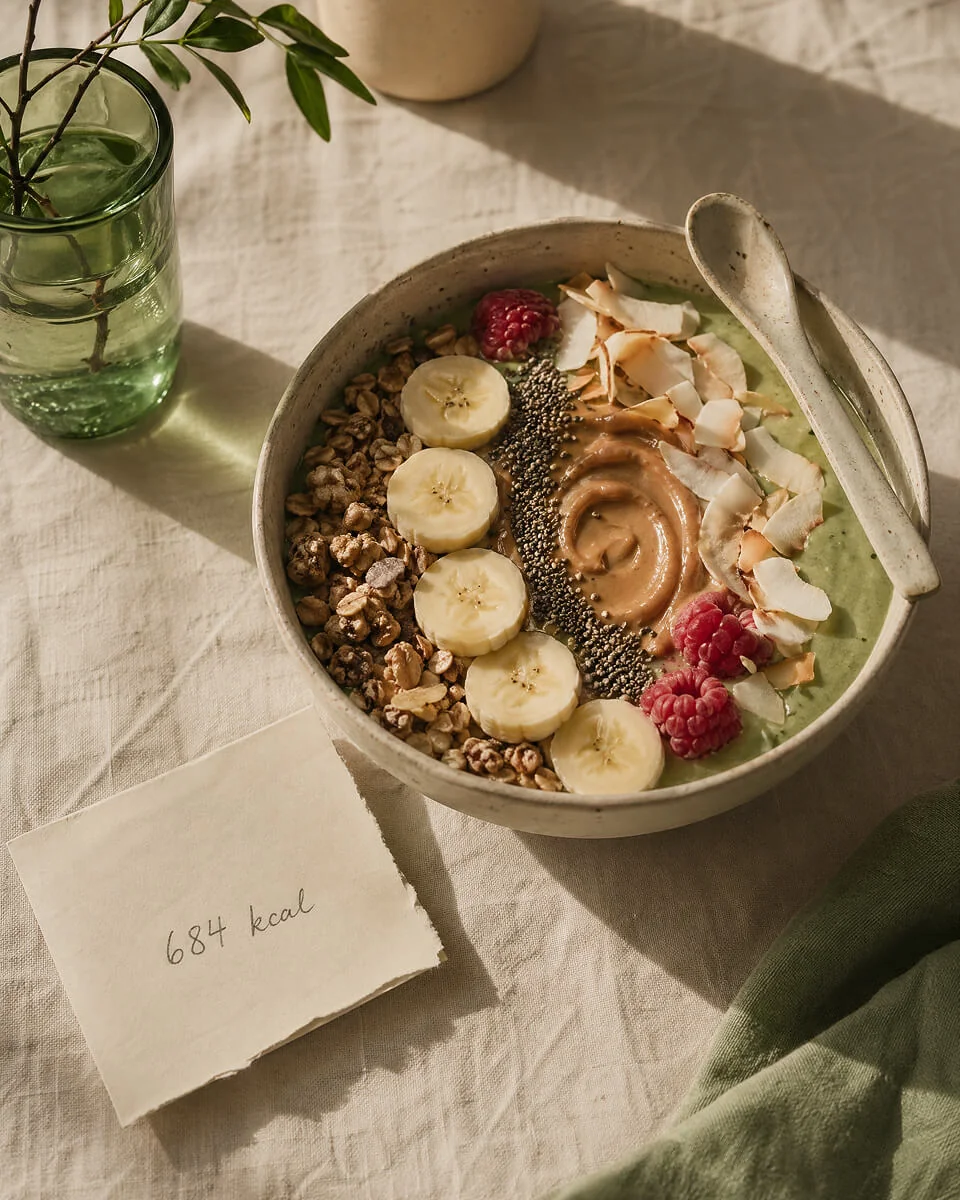 A green smoothie bowl topped with banana, granola, raspberries and toasted coconut, beside a hand-written 684 kcal note.