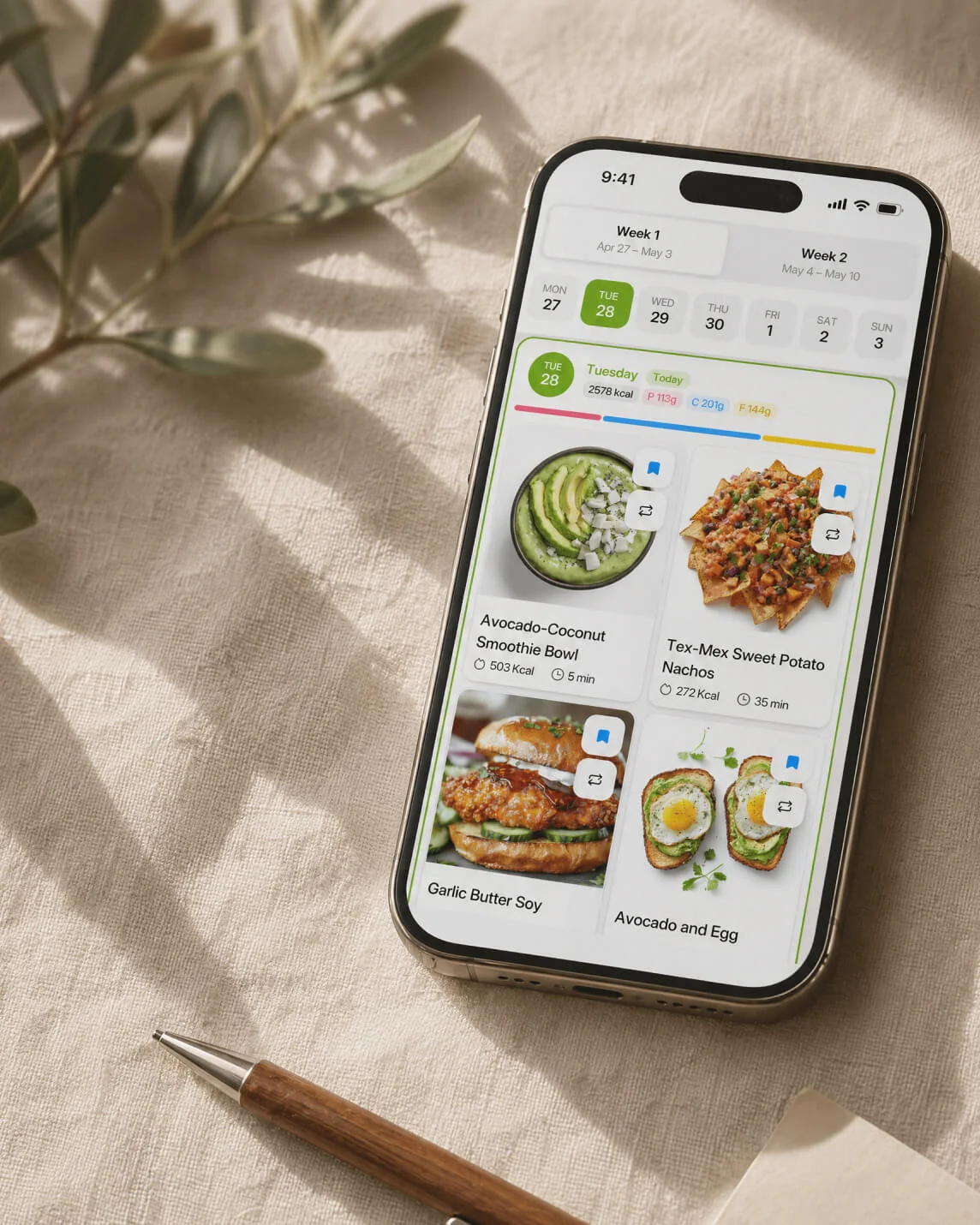 Nutriguide app weekly plan view — phone showing Tuesday's meals (avocado-coconut smoothie bowl, tex-mex sweet potato nachos, garlic butter soy, avocado and egg) on a linen surface beside a hand-written 'friday' note.