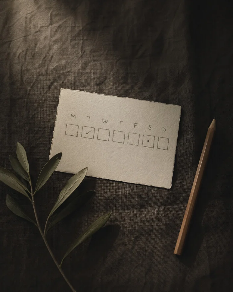 Hand-drawn weekly tracker on linen paper — M T W T F S S row of seven check-boxes, Tuesday ticked, Saturday marked with a quiet dot, beside an olive branch and a pencil. The week, observed.